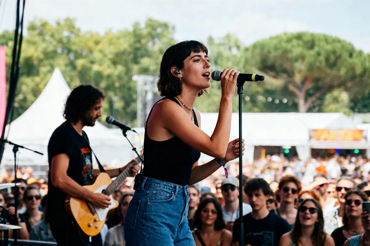 Chanteuse sur scène avec guitariste, micro, public en plein air, festival, journée ensoleillée, arbres en arrière-plan.