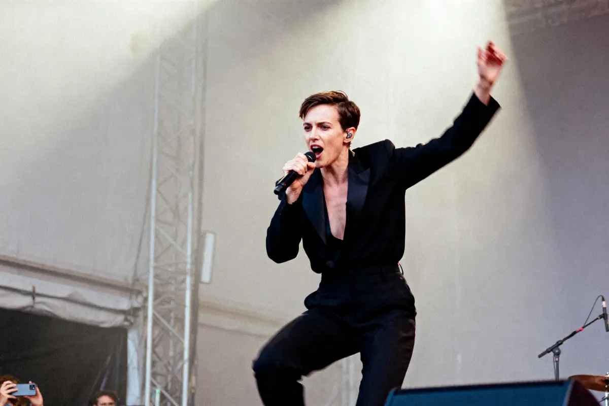 Chanteur en costume noir, micro à la main, sur scène lors d'un concert en extérieur, gesticulant énergiquement.