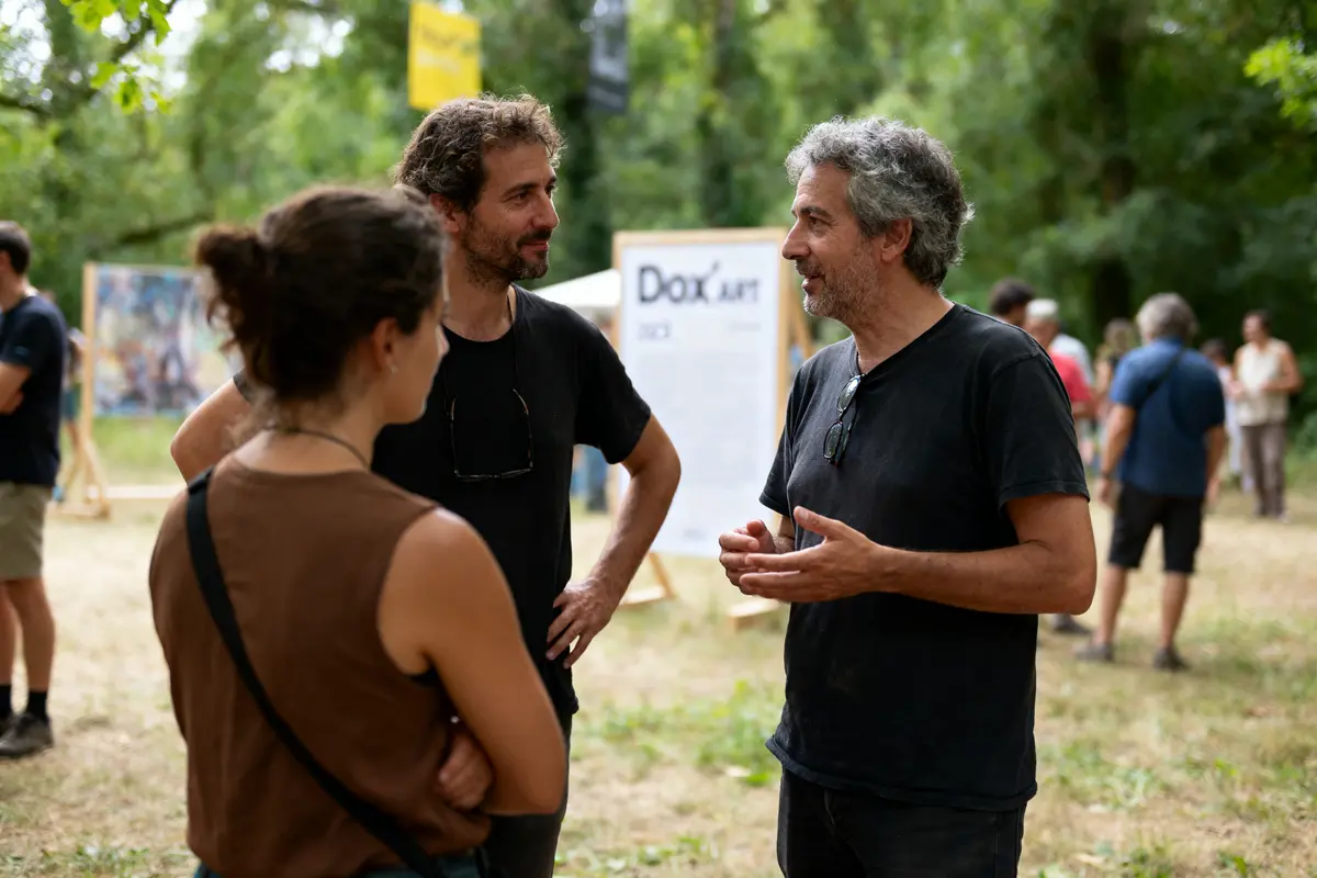 Trois personnes discutent en extérieur, avec des expositions en arrière-plan, lors d'un événement artistique en plein air.