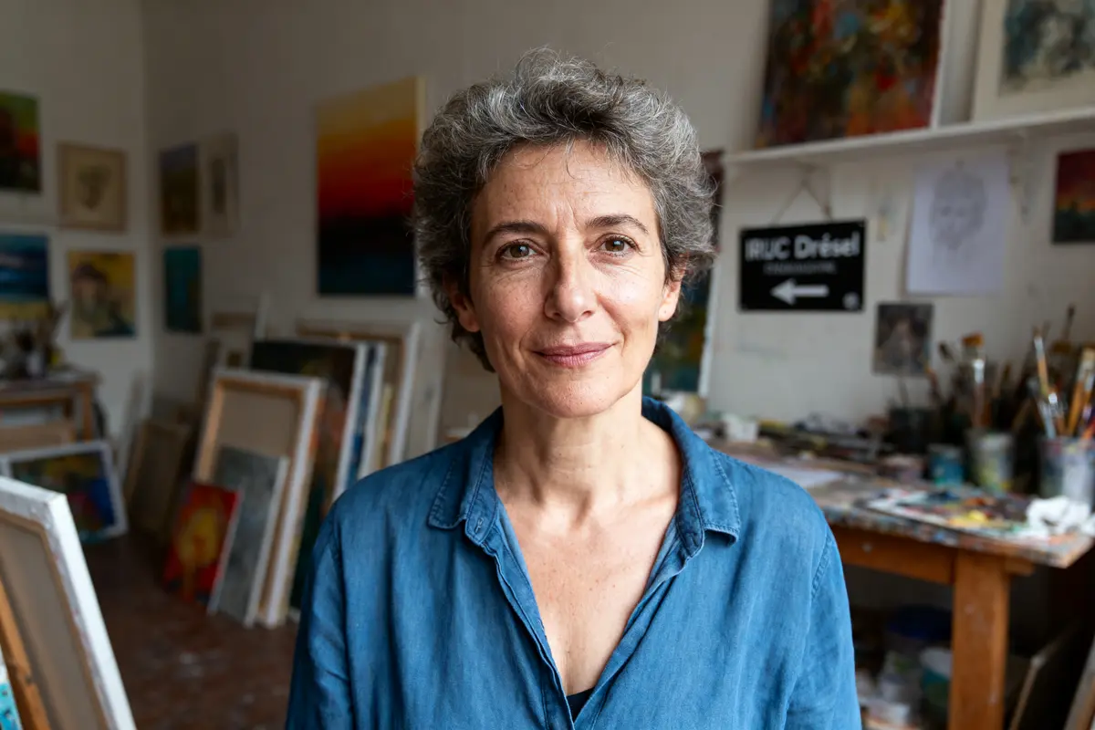 Une femme aux cheveux courts, chemise bleue, se tient dans un atelier d'art rempli de peintures.