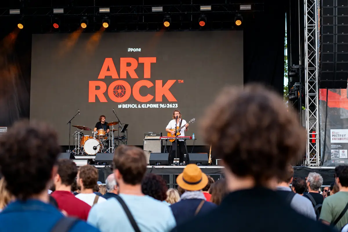 Concert à Art Rock, scène avec musiciens, guitare et batterie, public de dos, grande affiche en arrière-plan.