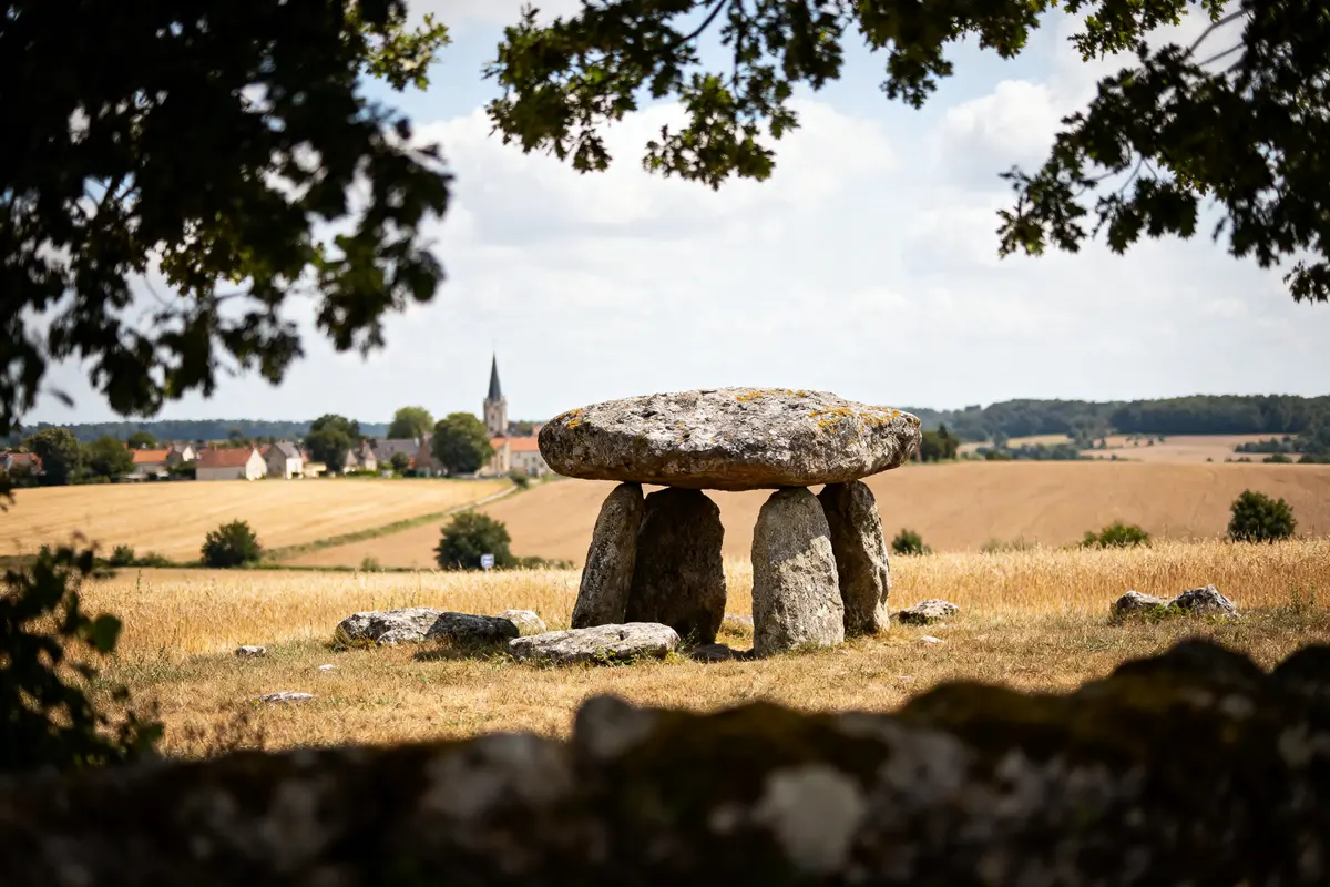 Un dolmen ancien dans un paysage champêtre avec village et clocher à l'horizon, sous ciel nuageux.