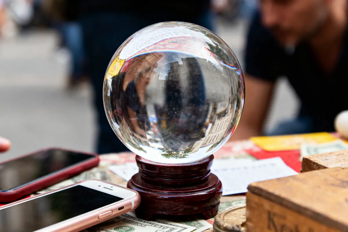 Boule de cristal sur support, entourée de smartphones et billets. Personne floue en arrière-plan.