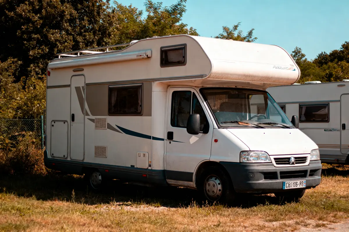 Camping-car blanc stationné dans un terrain herbeux, entouré d'arbres et de buissons.