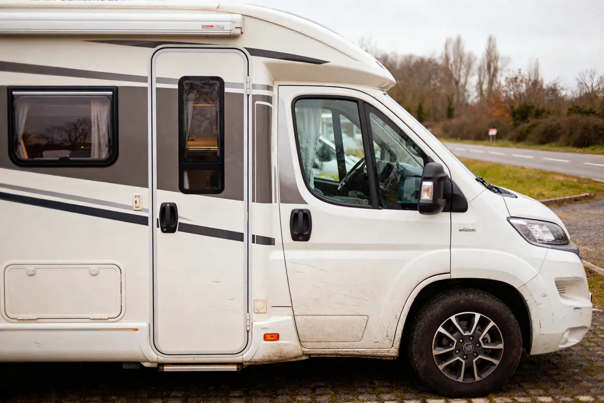 Camping-car blanc stationné sur une aire, route et arbres en arrière-plan, journée nuageuse.