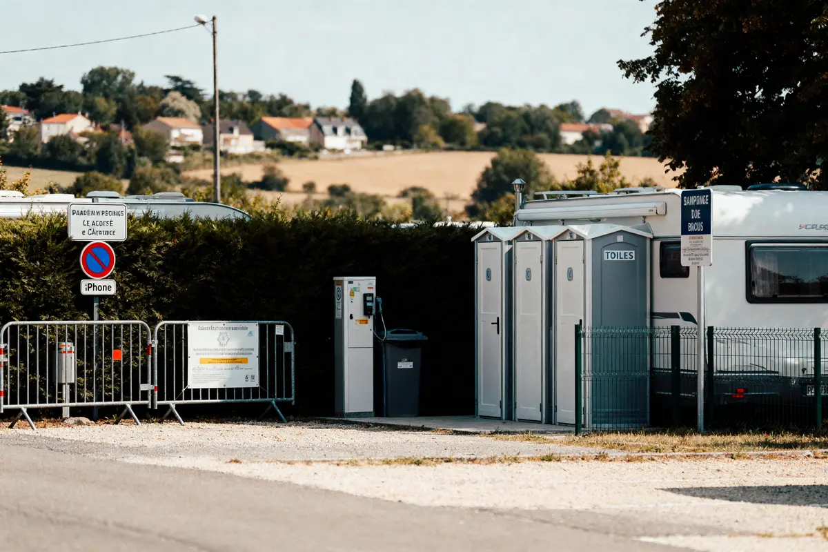 Toilettes portables et camping-car dans un parking, paysages de campagne en arrière-plan, panneaux de signalisation visibles.
