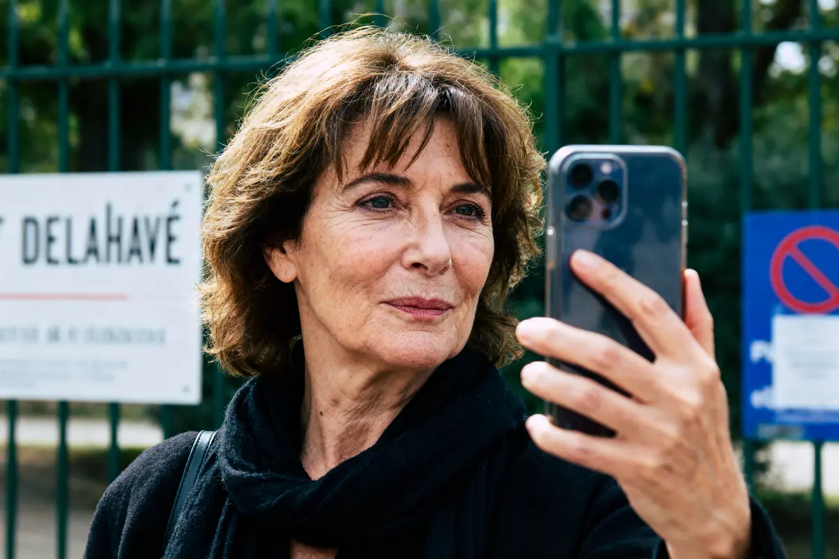 Femme prenant selfie devant panneau, extérieur, souriante, cheveux bruns, écharpe noire, portant un smartphone.